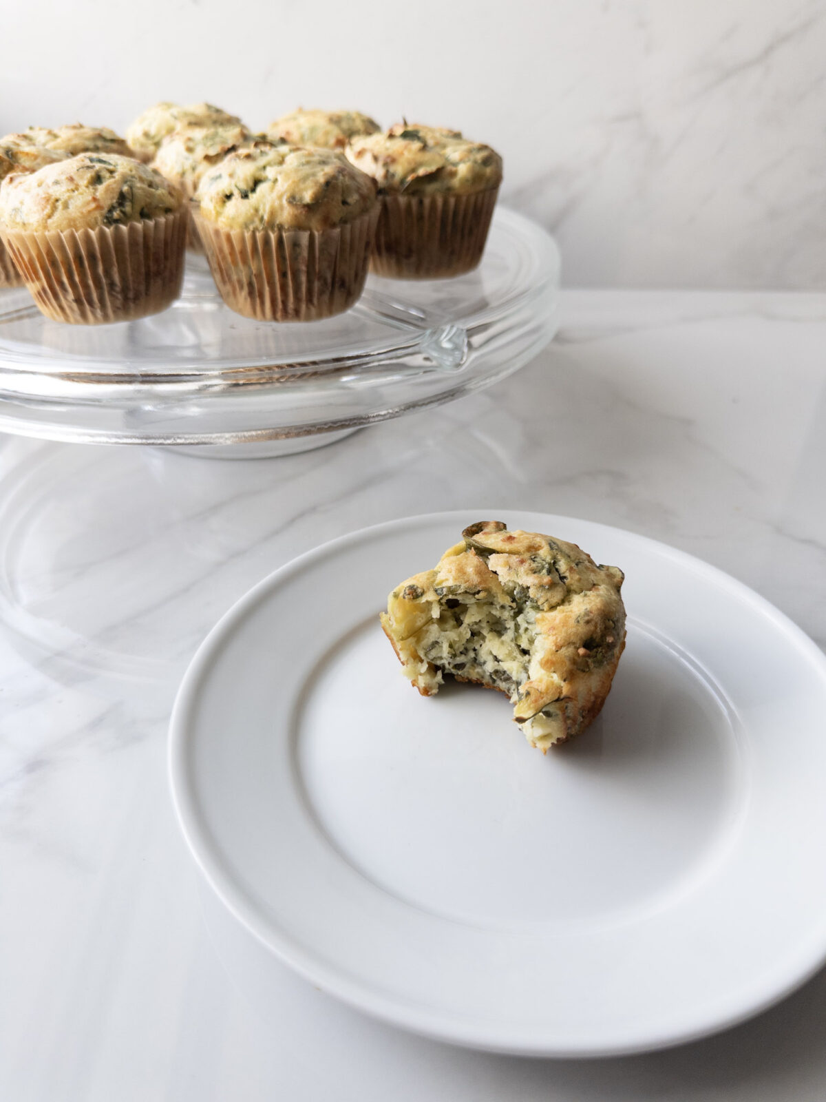 Savory Gluten-Free Spinach Feta Muffin on a plate with a bite taken out and a cake stand of muffins in the background