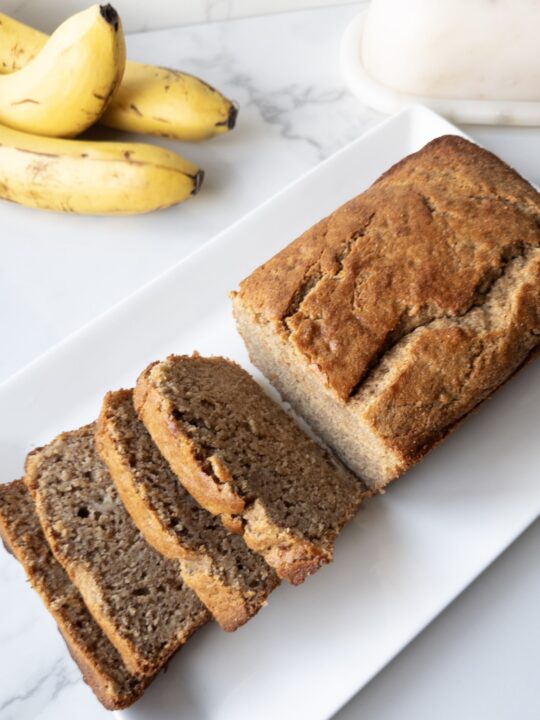 Sliced Fresh Milled Banana Bread sitting on a platter with bananas and a butter dish in the background