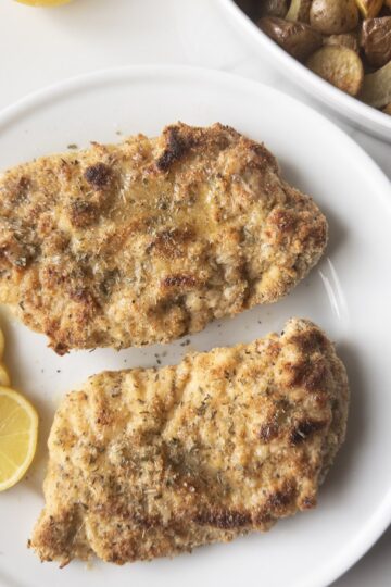 2 pieces of Gluten-Free Chicken Piccata on a plate with lemon slices and roasted potatoes in the background