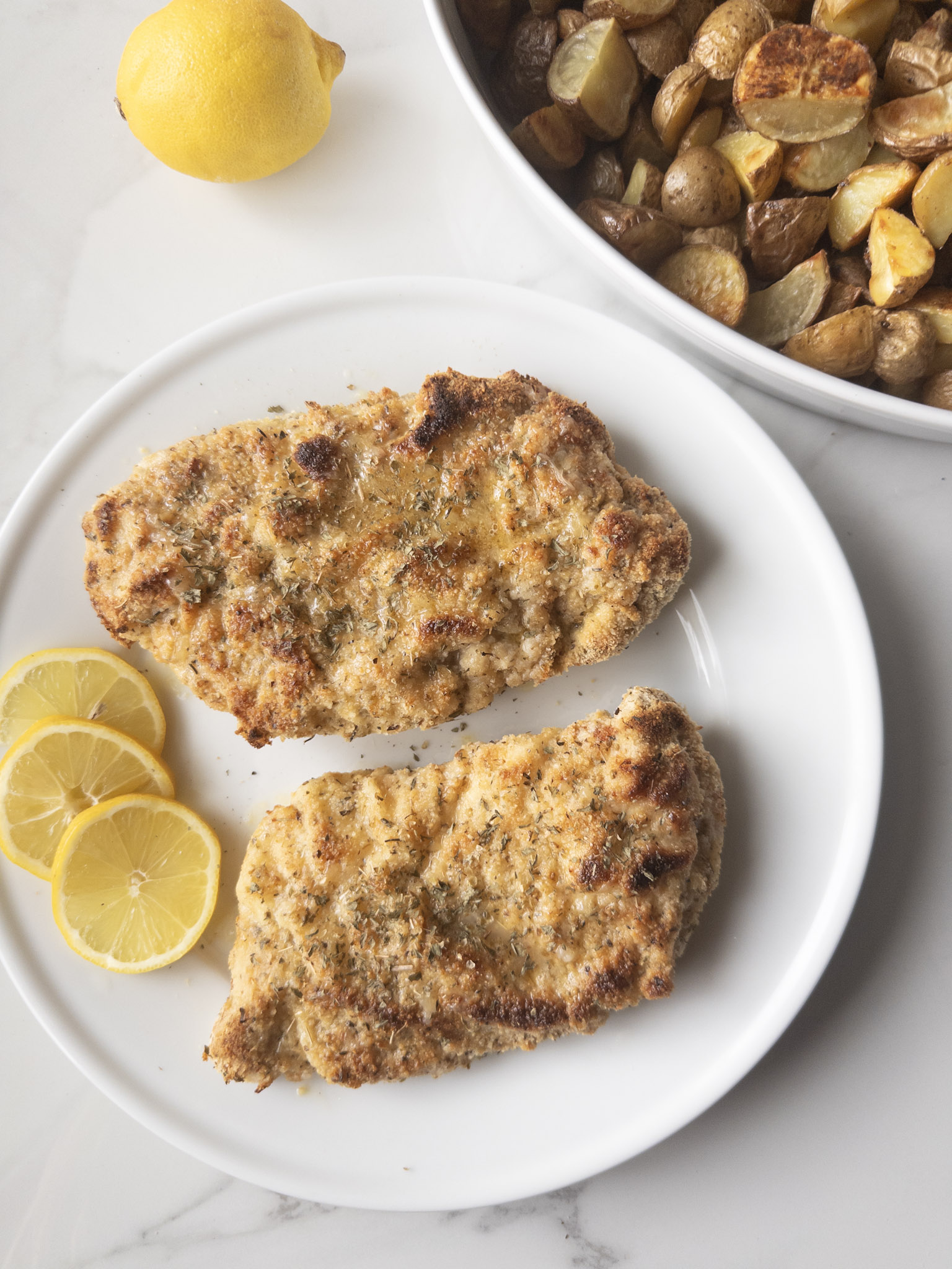 2 pieces of Gluten-Free Chicken Piccata on a plate with lemon slices and roasted potatoes in the background