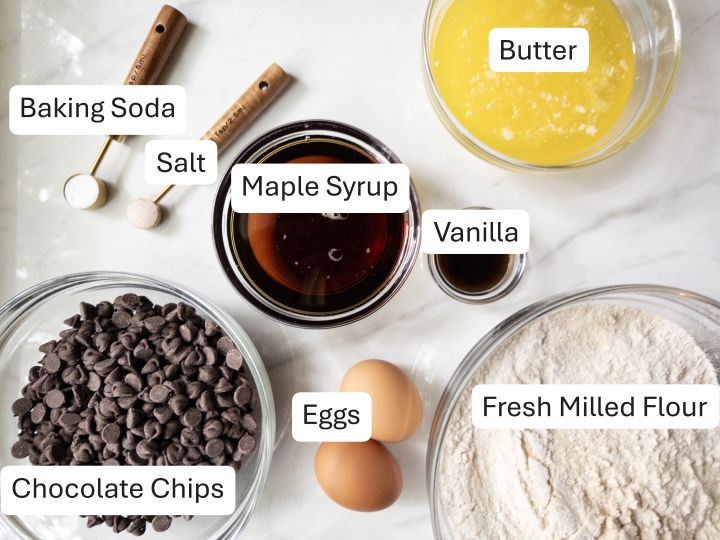 Ingredients for naturally sweetened fresh milled flour chocolate chip cookies, including fresh milled flour, maple syrup, melted butter, eggs, chocolate chips, baking soda, salt, and vanilla.