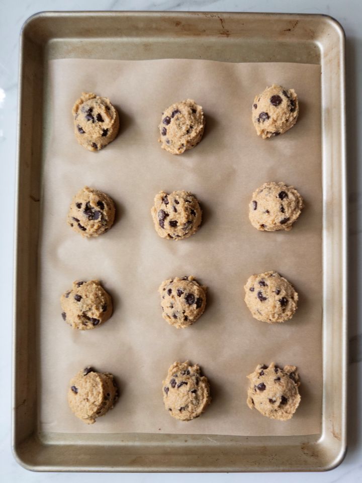Scooped balls of chocolate chip cookie dough arranged on a parchment-lined baking sheet, ready to bake.