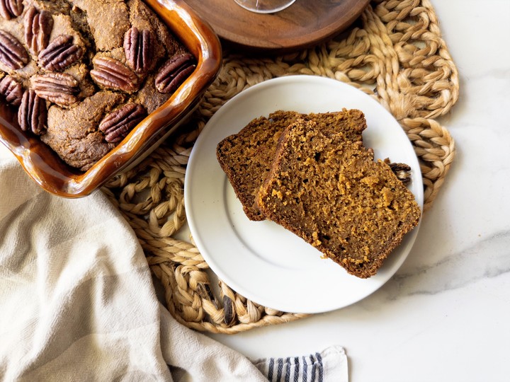 Overhead view of pumpkin bread topped with pecans in a baking dish, with two slices served on a white plate beside it