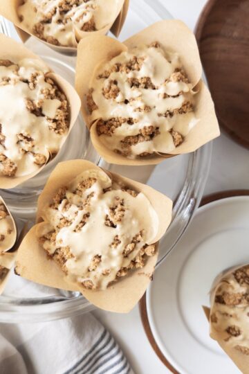 Overhead view of pumpkin muffins topped with crumbly streusel and drizzled with maple icing, arranged on a glass cake stand next to a glass of milk and a wooden bowl.