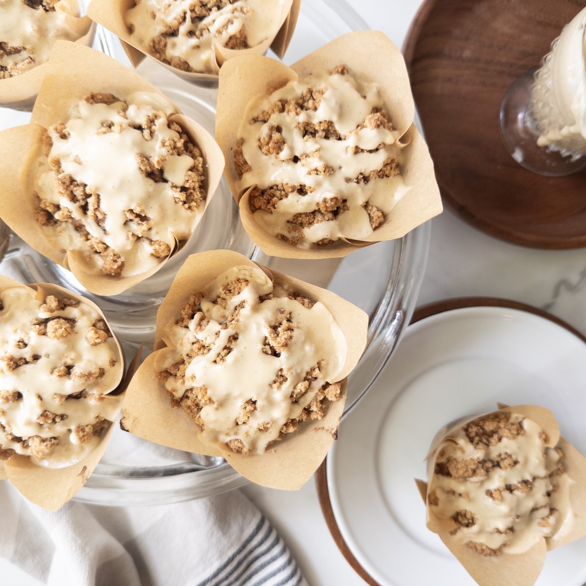 Overhead view of pumpkin muffins topped with crumbly streusel and drizzled with maple icing, arranged on a glass cake stand next to a glass of milk and a wooden bowl.