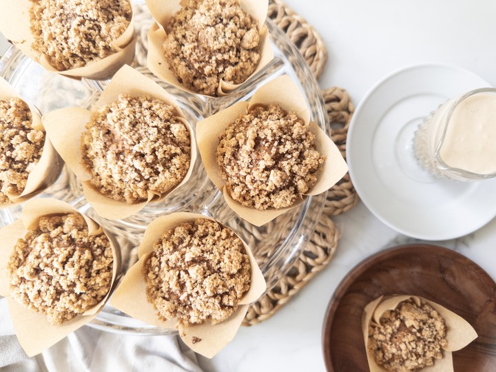 Overhead view of baked pumpkin muffins with golden streusel topping, arranged on a glass cake stand next to a glass of milk and a wooden bowl.