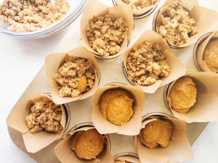 Overhead view of a muffin tin with unbaked pumpkin muffin batter in paper liners — some topped with crumbly streusel, others plain, with a bowl of extra streusel beside the pan.