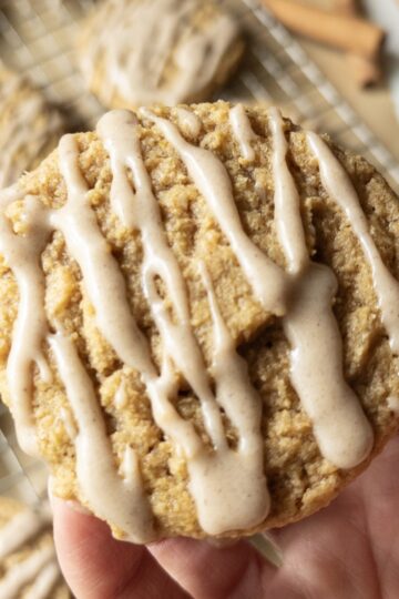 A hand holding a chewy pumpkin cookie drizzled with brown butter icing, with more cookies cooling on a wire rack and cinnamon sticks in the background.