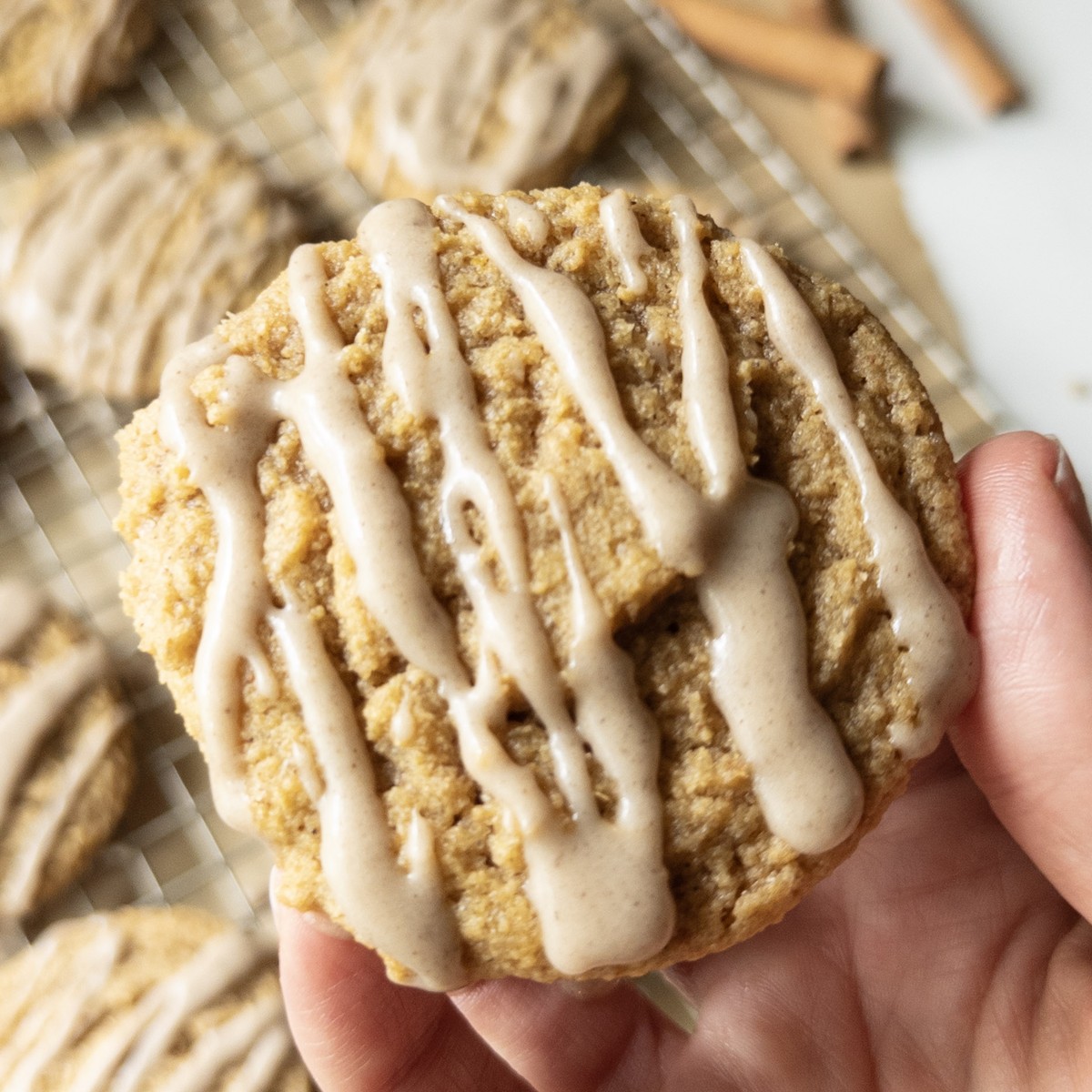 A hand holding a chewy pumpkin cookie drizzled with brown butter icing, with more cookies cooling on a wire rack and cinnamon sticks in the background.