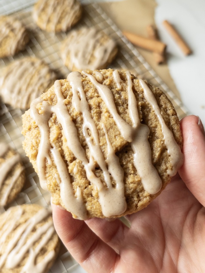 A hand holding a chewy pumpkin cookie drizzled with brown butter icing, with more iced cookies cooling on a wire rack in the background.