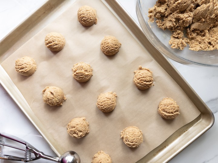 Cookie dough scooped into balls on a parchment-lined baking sheet, with the bowl of remaining dough beside it.