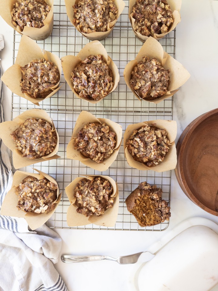 An overhead view of baked pumpkin muffins with pecan topping cooling on a wire rack next to a striped kitchen towel and wooden bowl, with one muffin cut in half.