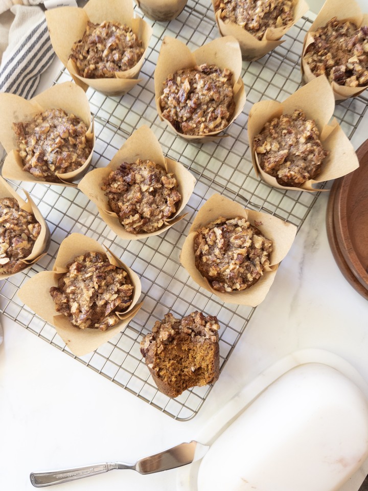 Baked pumpkin muffins with a maple pecan crunch topping cooling on a wire rack, with one muffin split open to show the moist crumb.
