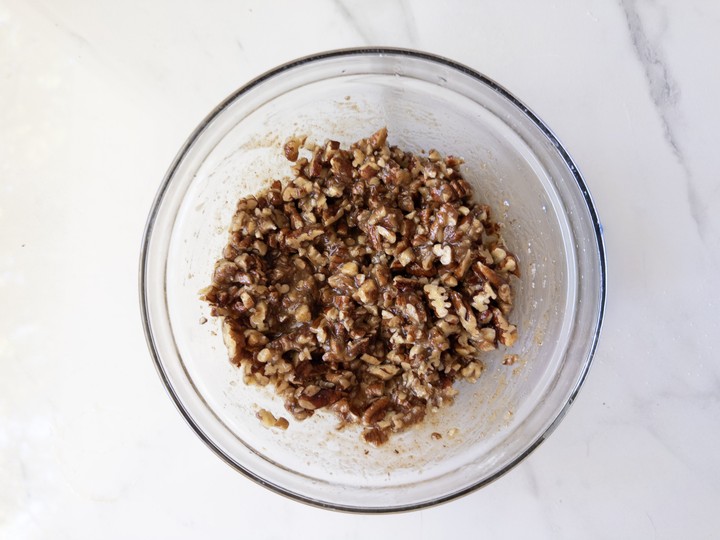 Chopped pecans mixed with melted butter, maple syrup, and vanilla extract in a glass bowl, just starting to clump together.