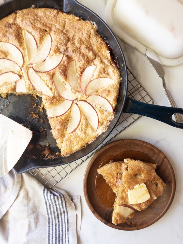 Overhead view of a puffed apple skillet pancake studded with apple slices, with a sliced serving topped with butter and syrup on a plate beside it.