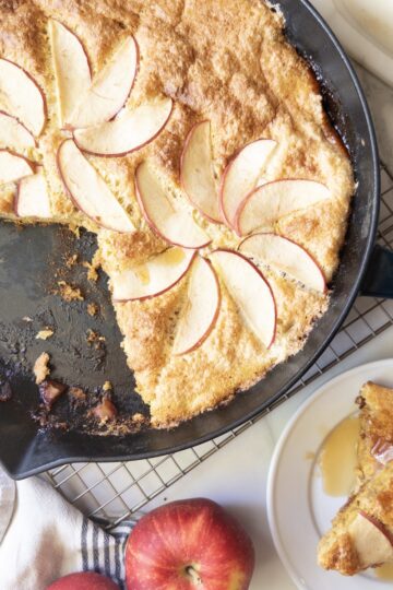 Overhead view of a puffed apple skillet pancake in a cast iron skillet, topped with apple slices, with a sliced serving on a plate with butter and syrup.