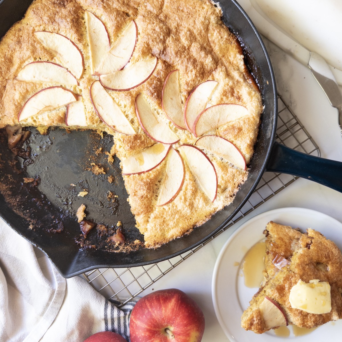 Overhead view of a puffed apple skillet pancake in a cast iron skillet, topped with apple slices, with a sliced serving on a plate with butter and syrup.