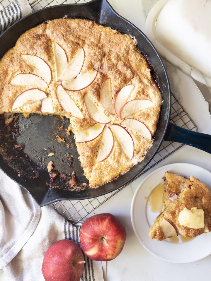 Overhead view of a puffed apple skillet pancake studded with apple slices, with a sliced serving topped with butter and syrup on a plate beside it.