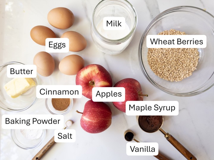 Labeled ingredients for puffed apple skillet pancake on a marble counter, including wheat berries, eggs, apples, butter, milk, cinnamon, baking powder, salt, maple syrup, and vanilla.