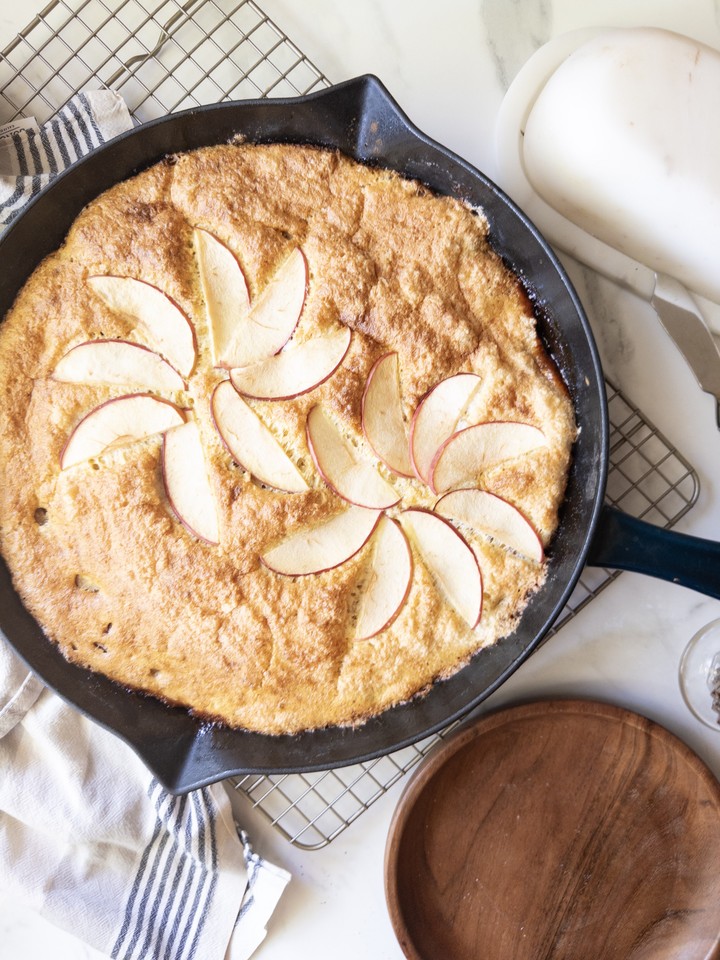 Overhead view of the baked puffed apple pancake in a cast iron skillet, topped with a fan of apple slices, cooling on a wire rack.