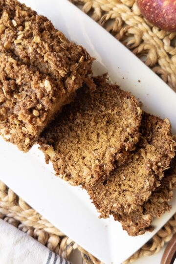 Sliced apple crisp bread with a cinnamon oat crumb topping on a white rectangular plate beside a red apple.