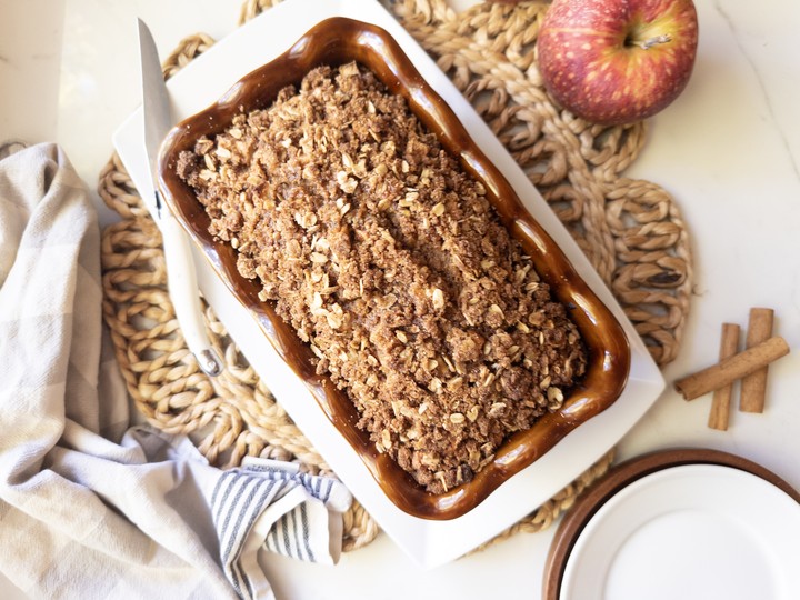 Baked apple crisp bread topped with an oat crumble in a brown loaf pan on a white plate, with red apples and cinnamon sticks nearby.