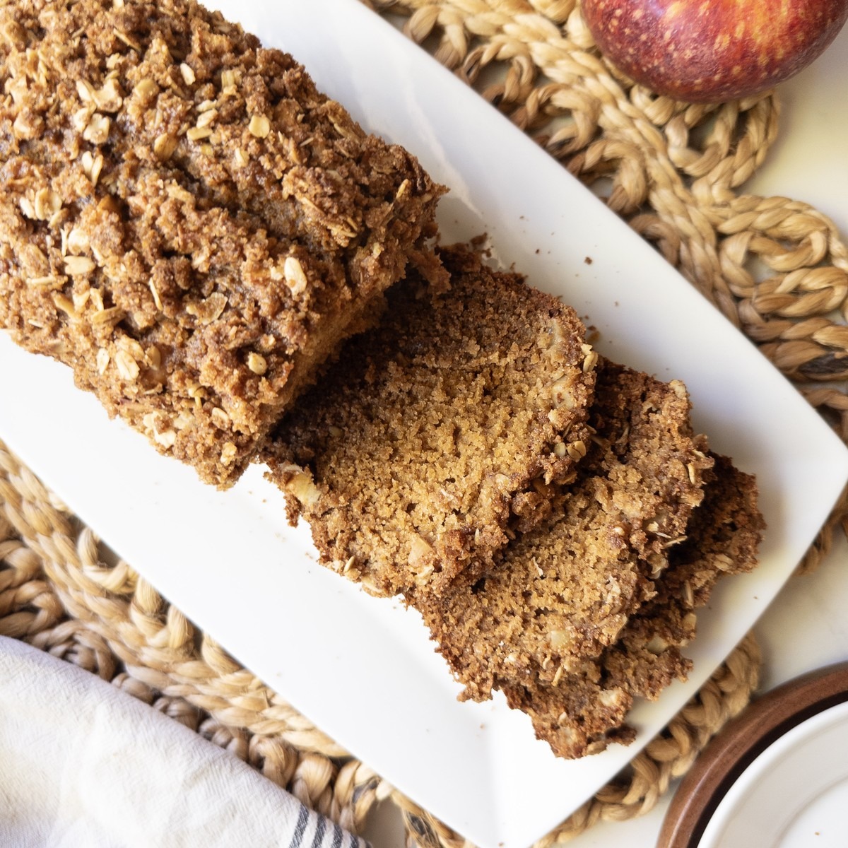 Sliced apple crisp bread with a cinnamon oat crumb topping on a white rectangular plate beside a red apple.