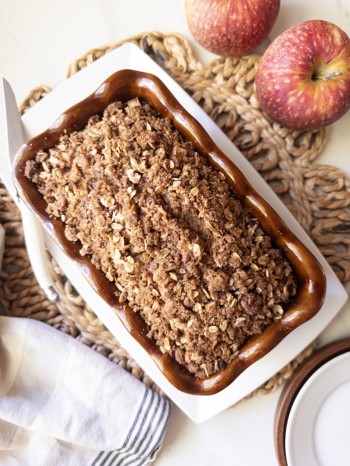 Overhead view of a baked apple crisp bread in a brown loaf pan on a white plate with two red apples beside it.