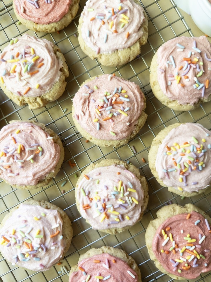 Close-up of soft sugar cookies topped with pink buttercream and rainbow sprinkles on a wire cooling rack.