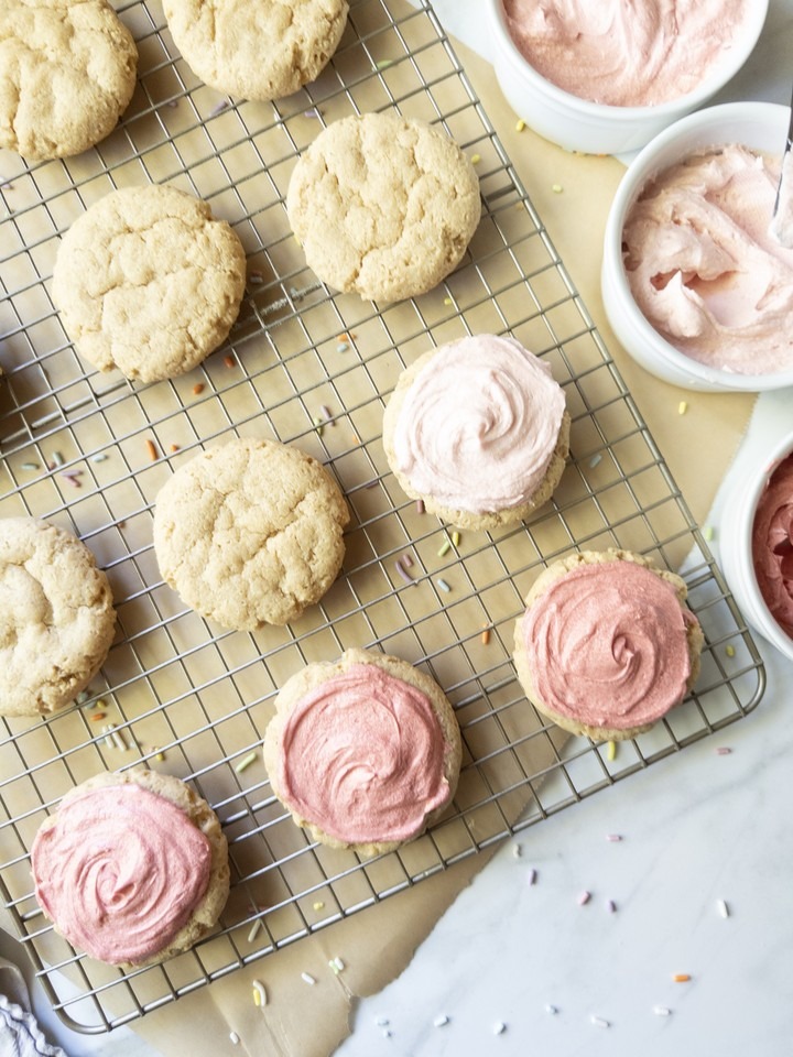 Sugar cookies on a wire cooling rack, some plain and some topped with light and dark pink buttercream, next to bowls of frosting.