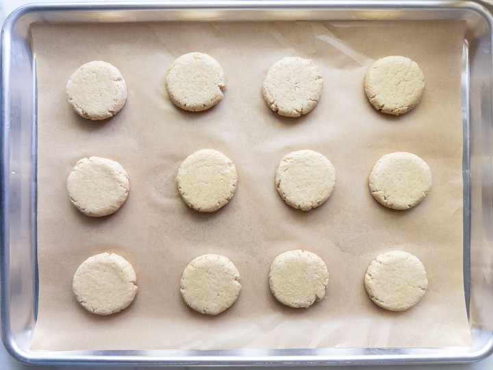 Twelve flattened rounds of sugar cookie dough on a parchment-lined cookie sheet.
