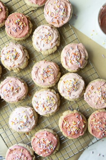 Overhead view of soft frosted sugar cookies with pink buttercream and rainbow sprinkles on a wire cooling rack, with bowls of pink frosting nearby.