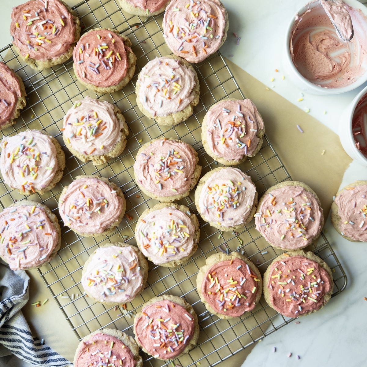 Overhead view of soft frosted sugar cookies with pink buttercream and rainbow sprinkles on a wire cooling rack, with bowls of pink frosting nearby.