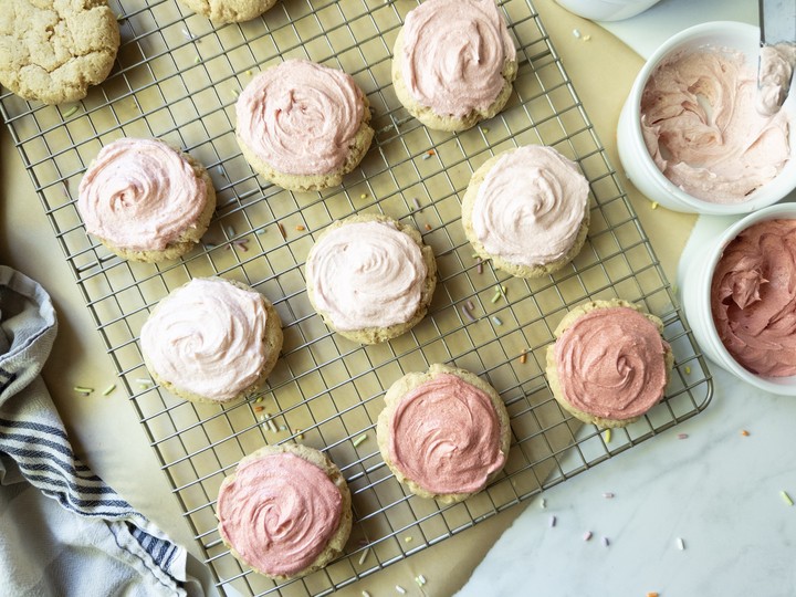 Soft frosted sugar cookies with pink buttercream on a wire cooling rack, with bowls of pink frosting beside them.
